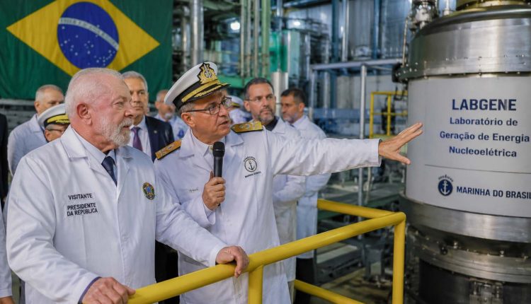 Presidente Lula acompanha explicações de integrante da Marinha do Brasil durante visita a instalação industrial do programa nuclear brasileiro, com equipamentos do Labgene e a bandeira do Brasil ao fundo.